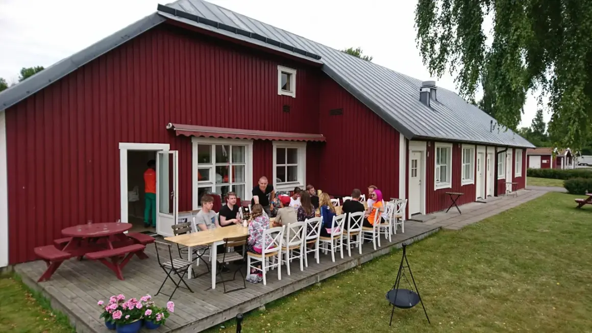 Gästerna njuter av en måltid utomhus vid Sjötorp vandrarhem. En grupp vänner samlas runt borden på altanen framför den röda byggnaden.