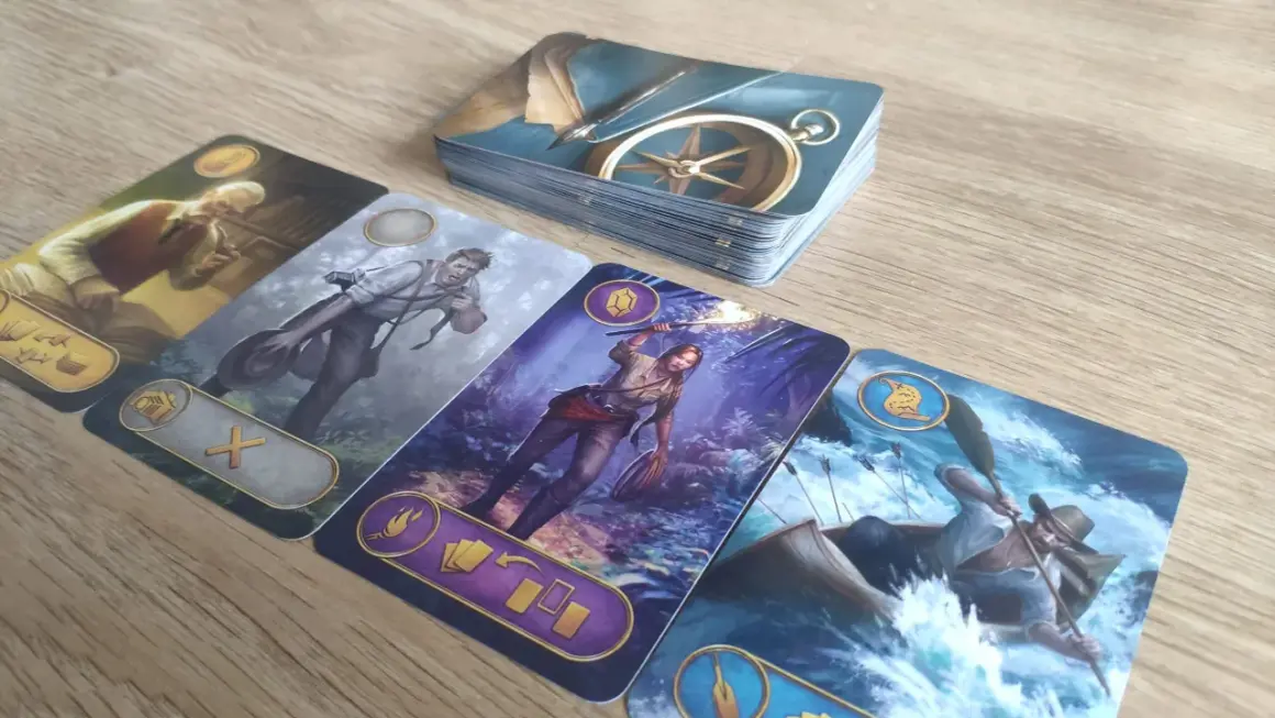 Na stole leżą karty z gry ryzykanci. Widoczny jest stos kart z kompasem, a obok cztery karty przedstawiające postacie: starszego pana z fajką, mężczyznę z aparatem, kobietę z pochodnią i mężczyznę w łodzi na rwącej rzece.
