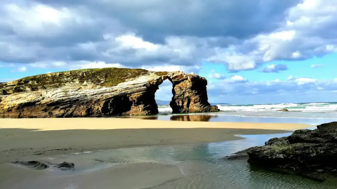 Arco natural en las playas de Galicia, con arena dorada, mar azul y cielo nublado.