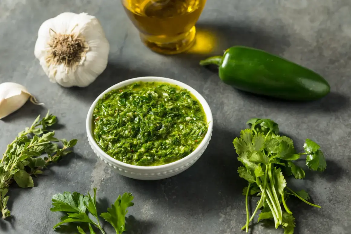 Miseczka pełna aromatycznego sosu chimichurri, obok świeża kolendra, natka pietruszki, czosnek, papryczka jalapeno i oliwa.