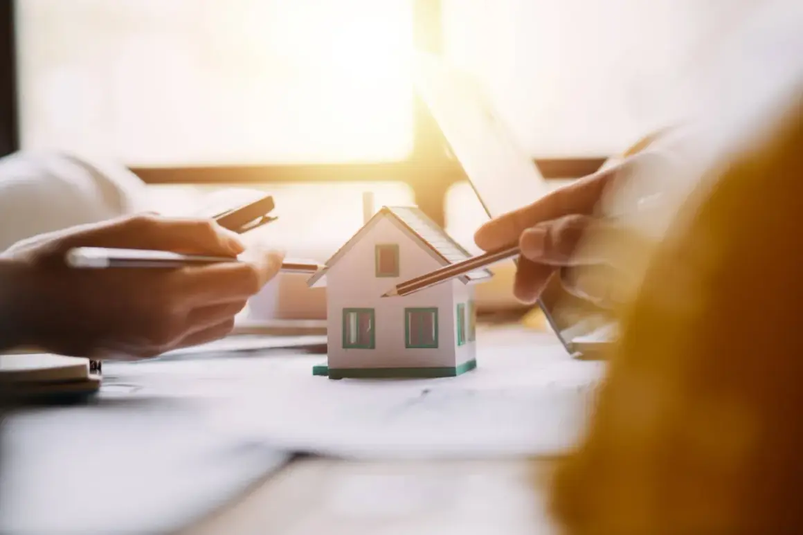 Mani che studiano un modello di casa, con matite e calcolatrice. Si discute il surroga mutuo significato per un nuovo acquisto immobiliare.