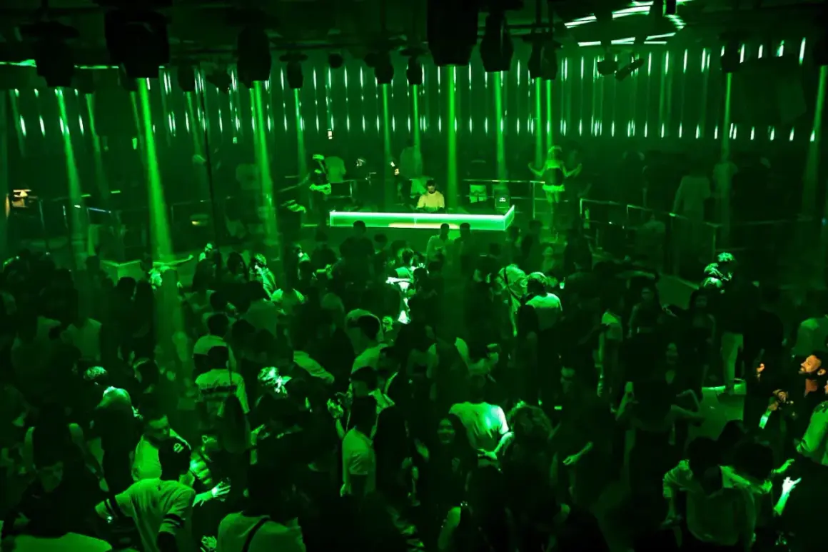 A crowded nightclub with green lights and a DJ booth.