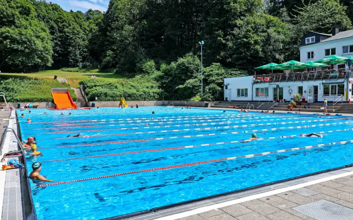 Ein sonniger Tag im Freibad Neuenhof. Menschen schwimmen in den Bahnen des großen Pools, während im Hintergrund grüne Bäume und ein Gebäude mit Sonnenschirmen zu sehen sind.