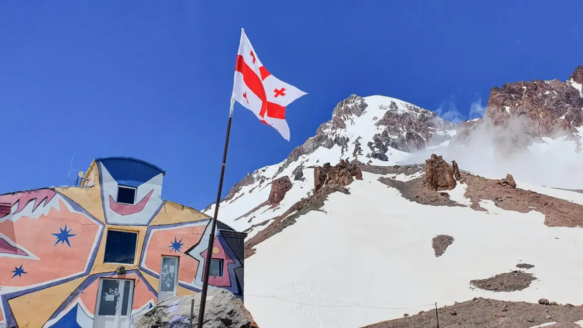 Kolorowy budynek i gruzińska flaga na tle ośnieżonych szczytów. Kazbek wysokość robi wrażenie.