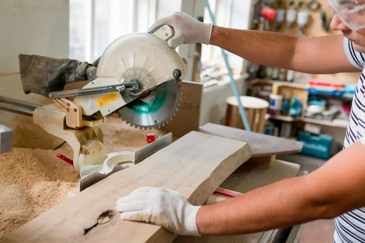 Un artisan utilise une scie à onglet pour couper une planche de bois. Il tient la pièce avec des gants, prêt à réaliser une coupe précise.