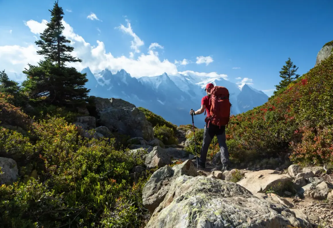 Randonneur avec sac à dos rouge, bâtons de marche, sur un sentier rocheux. Montagnes enneigées en arrière-plan. Combien de jours de marche pour atteindre ce sommet ? gr5.