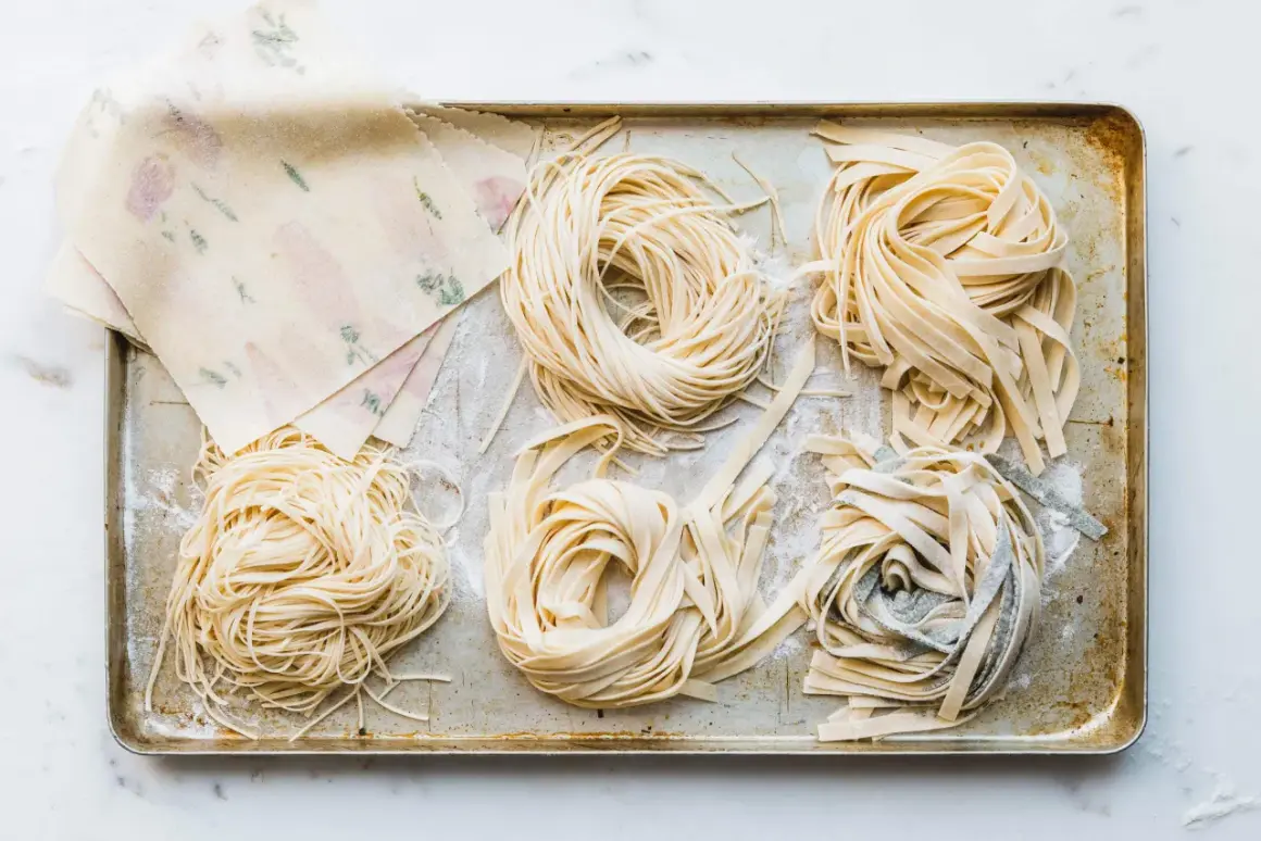 Frische Nudeln in verschiedenen Formen liegen auf einem Blech. Ein Stapel mit Blumenmuster verzierter Teigblätter liegt daneben. Perfekt für alle, die Nudeln selber machen wollen.