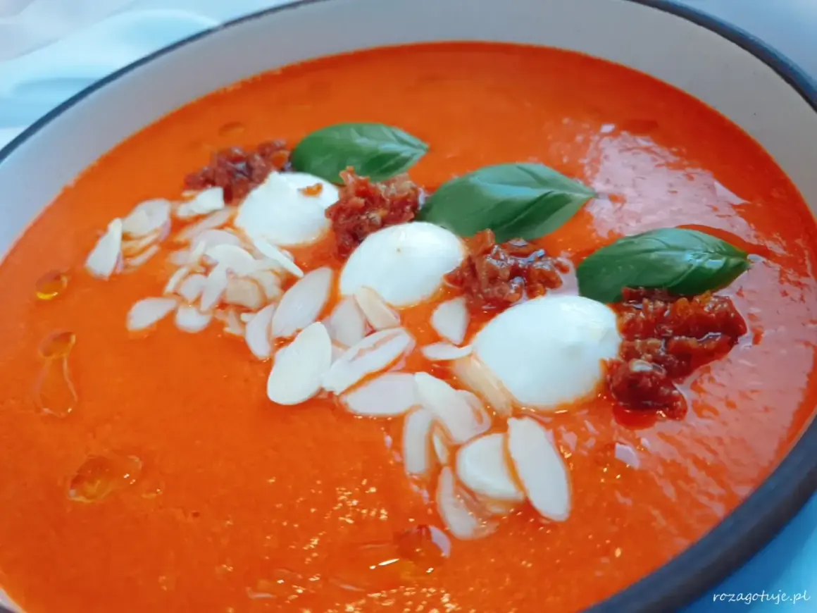 Aksamitna zupa z pieczonej papryki, ozdobiona kulkami mozzarelli, suszonymi pomidorami, płatkami migdałów i listkami bazylii.
