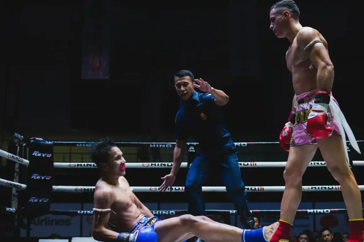 Domare stoppar en match i muy thai. En fighter ligger på mattan, den andra står redo.