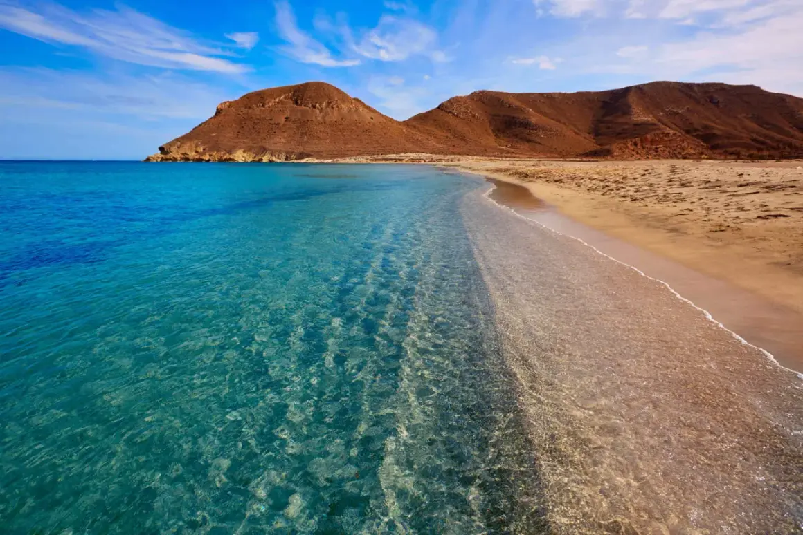 Aguas cristalinas acarician la arena dorada de las playas vírgenes de Almería, con montañas áridas de fondo bajo un cielo azul.