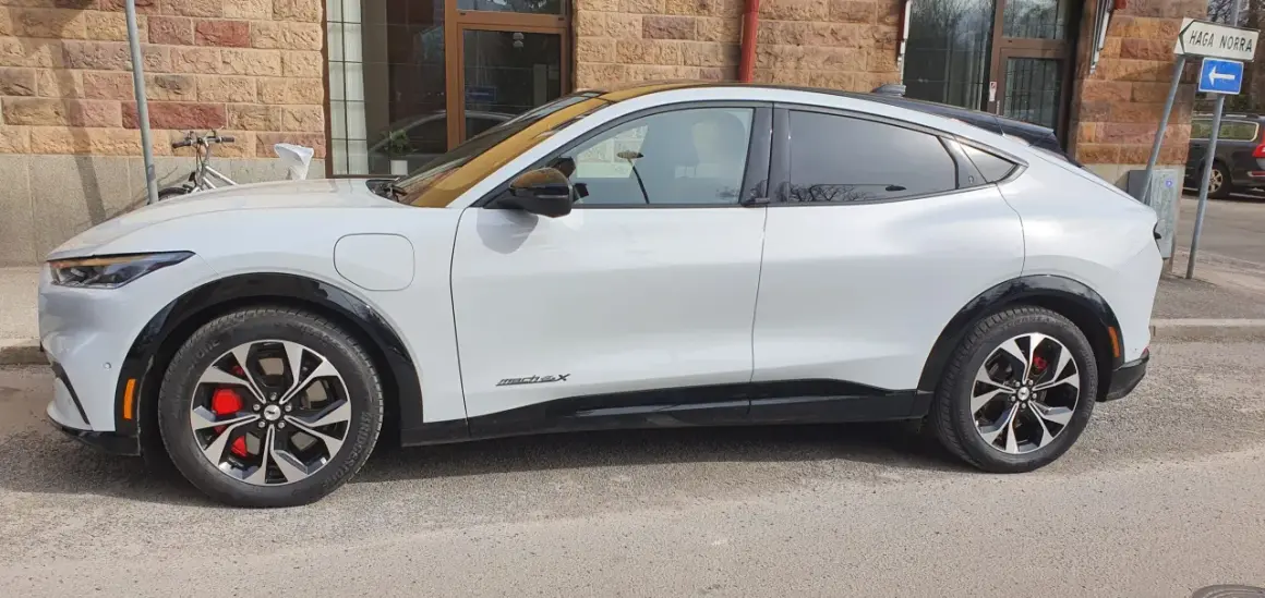 Vit Ford Mustang Mach-E står parkerad utanför en tegelbyggnad. En cykel syns i bakgrunden. Detta är en ford mustang mach-e test.