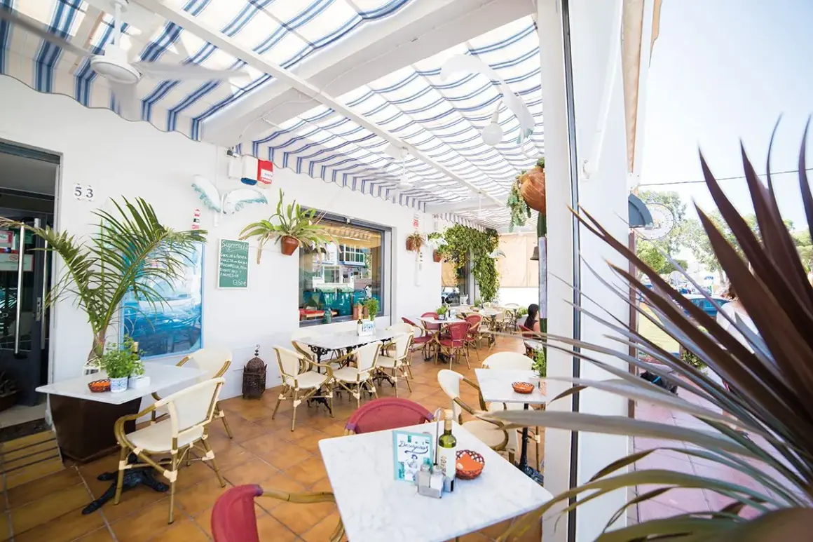 Terraza de restaurante con mesas y sillas, toldos a rayas y vegetación. Un cartel anuncia "il brigantino san jose".