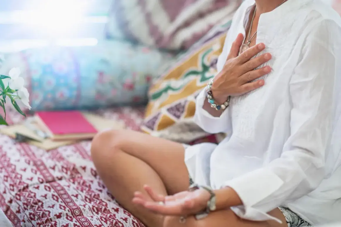 Frau in weißer Kleidung praktiziert buddhistische Herzmeditation, Hände auf der Brust und im Schoß, umgeben von Kissen und Büchern.