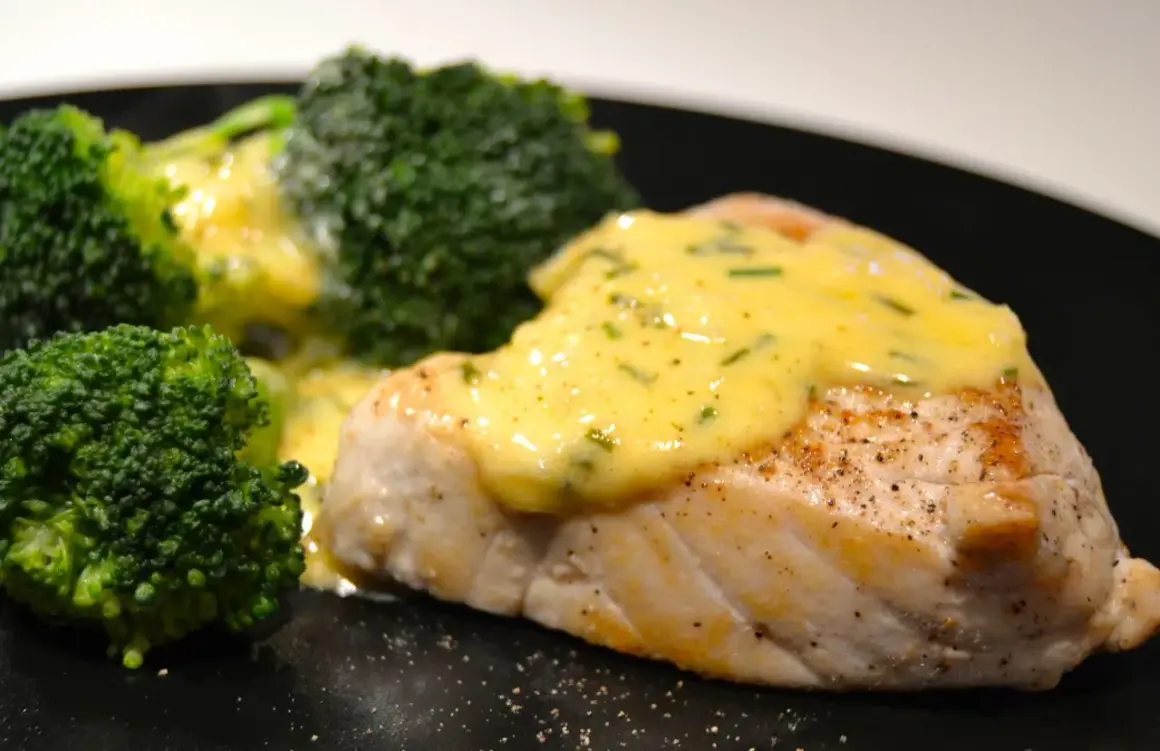 En saftig fiskfilé med krämig sås till tonfisk och gröna broccolibuketter på en svart tallrik.