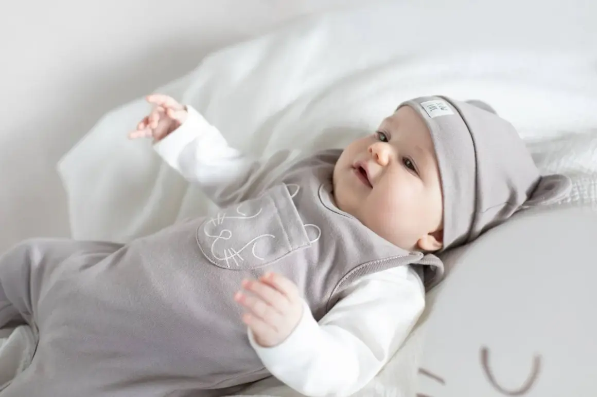 Uroczy niemowlak w szarym śpioszkowym kombinezonie z uszkami i haftem kotka, leżący na białym tle.