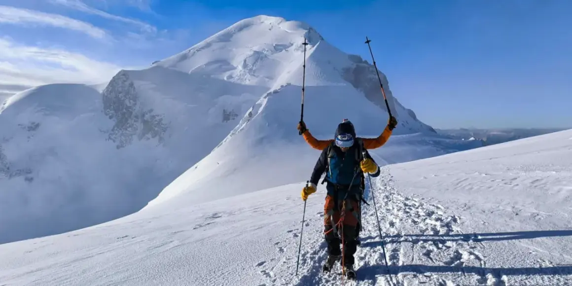Un alpiniste victorieux lève ses bâtons sur fond de Mont Blanc. L'ascension est réussie, la neige brille sous le ciel bleu.