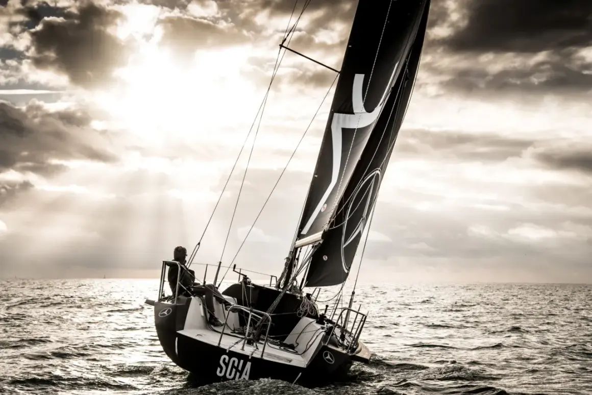 Vela nera con logo, un uomo a bordo di una barca a vela che naviga in mare con un cielo drammatico. L'imbarcazione mostra un angolo di deriva accentuato.