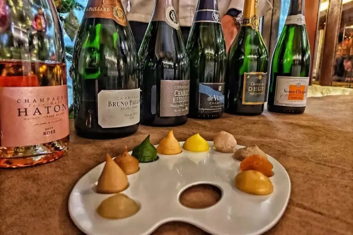 Bottiglie di champagne, tra cui Veuve Clicquot e Bruno Paillard, disposte su un tavolo con una tavolozza da pittore piena di dolcetti colorati, un'ode al gusto e all'arte del cote de bar.