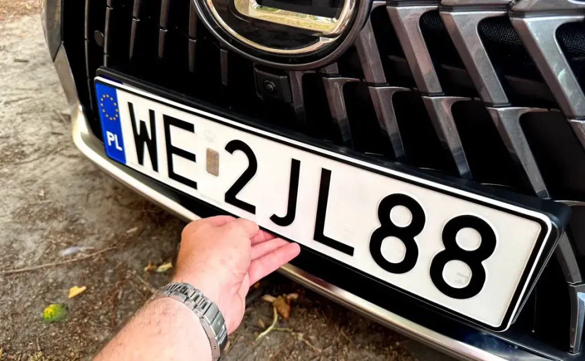 Dłoń poprawia tablicę rejestracyjną auta. Wartość samochodu powyżej 150 tys. zł, widoczna na grillu i zderzaku.