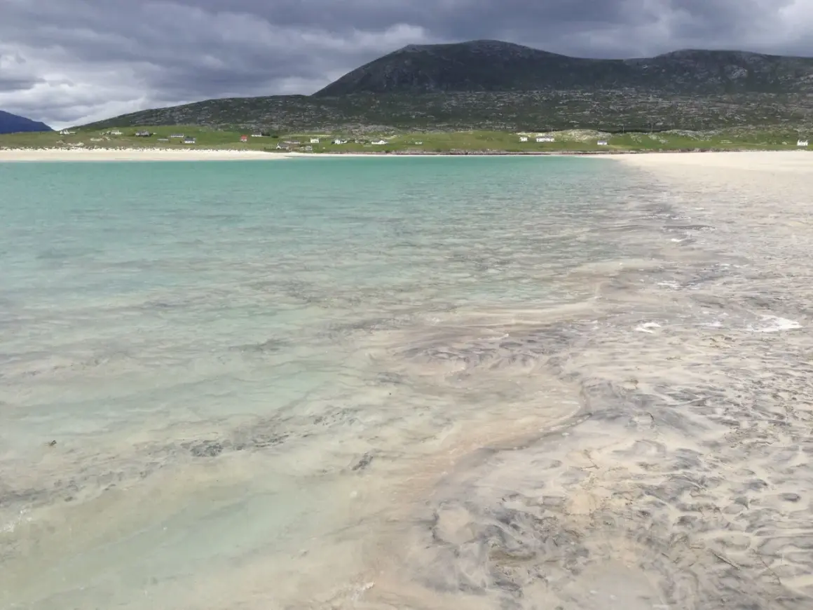 Vackert turkost vatten möter en sandstrand med mönster från tidvattnet. Hus och en grön kulle syns i bakgrunden på Yttre Hebriderna.