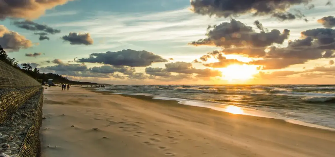 Zachód słońca nad Bałtykiem, plaża i falochron. W oddali widać ludzi spacerujących i kitesurferów. To idealne miejsce na podziwianie atrakcji Jastrzębiej Góry i okolic.