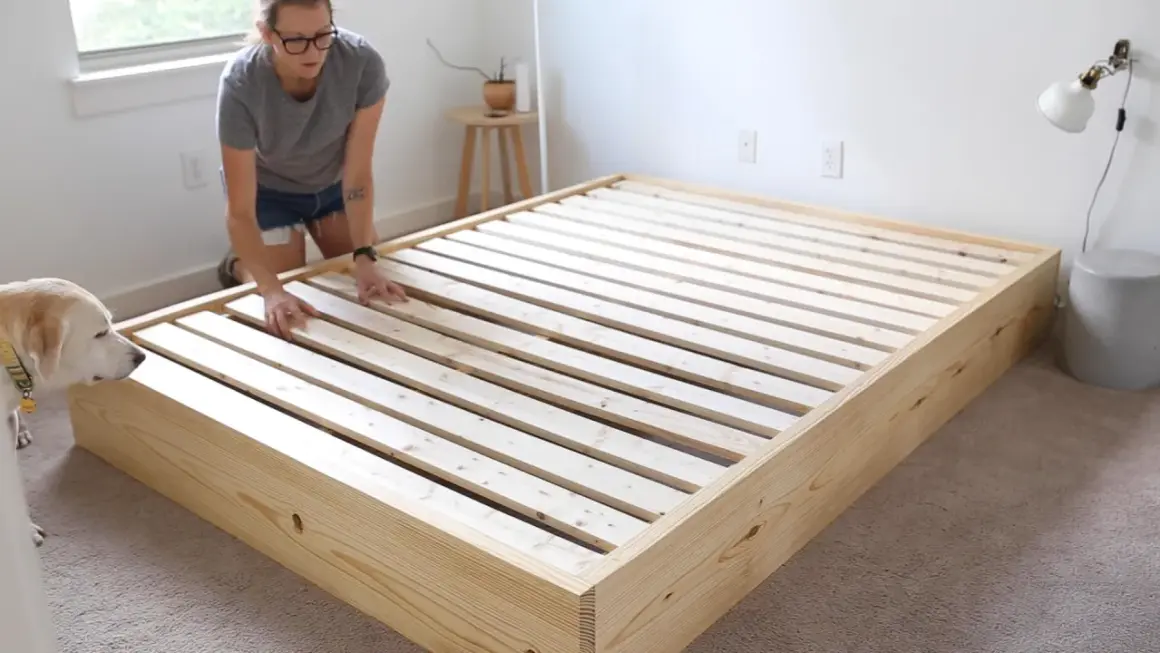 A woman and her dog inspect a newly built DIY platform bed frame made of light wood.
