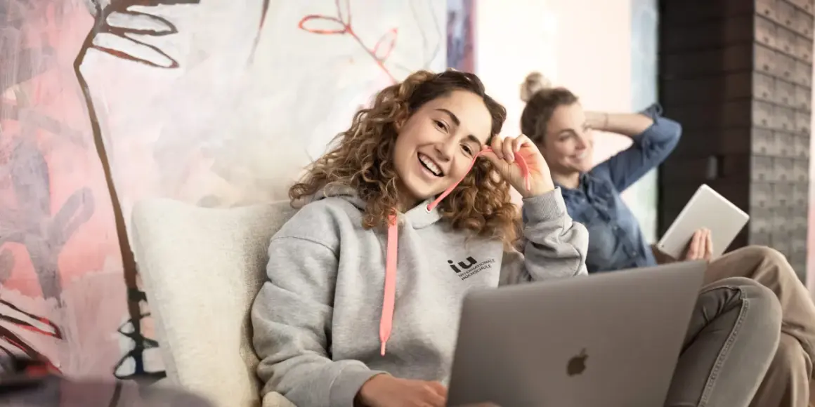 Zwei junge Frauen lernen entspannt. Eine trägt einen Hoodie mit dem Logo der IU Internationale Hochschule, die andere hält ein Tablet.