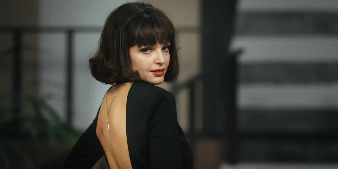 Elegancka Maria Dębska w czarnej sukni z głębokim wycięciem na plecach, zmysłowo odwraca głowę.