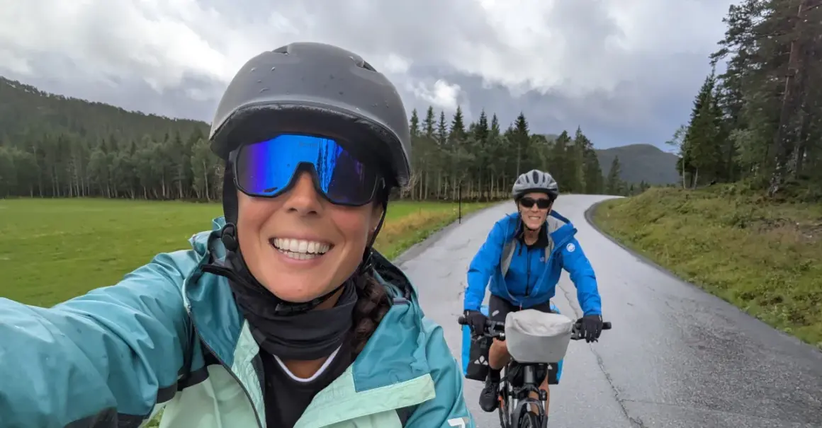 Dos ciclistas disfrutan de la ruta EuroVelo 6, con un paisaje verde y montañoso bajo un cielo nublado.
