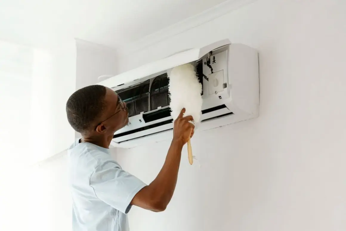 Un homme nettoie son climatiseur avec un plumeau. Un entretien régulier est essentiel pour savoir entretien climatisation tous les combien.