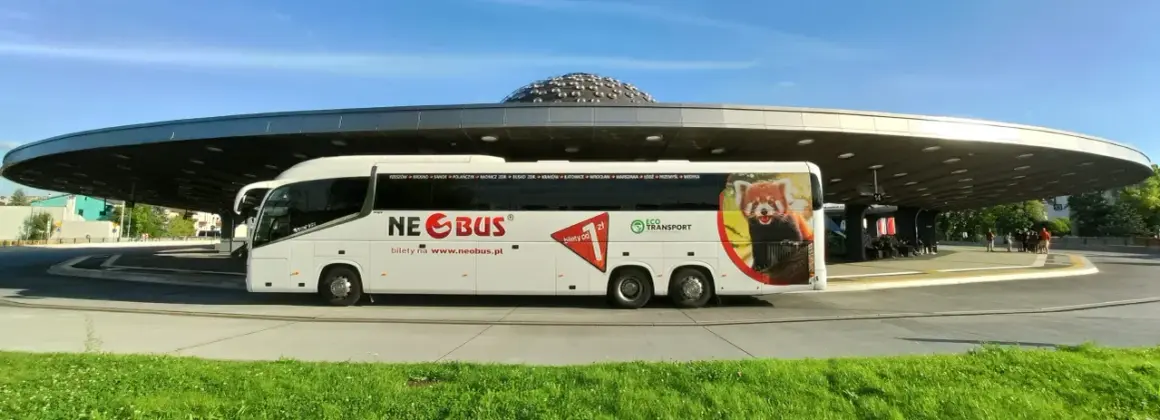 Biały autokar Neobus z wizerunkiem pandki rudej na tle nowoczesnego dworca. Sprawdź neobus ceny biletów na www.neobus.pl.