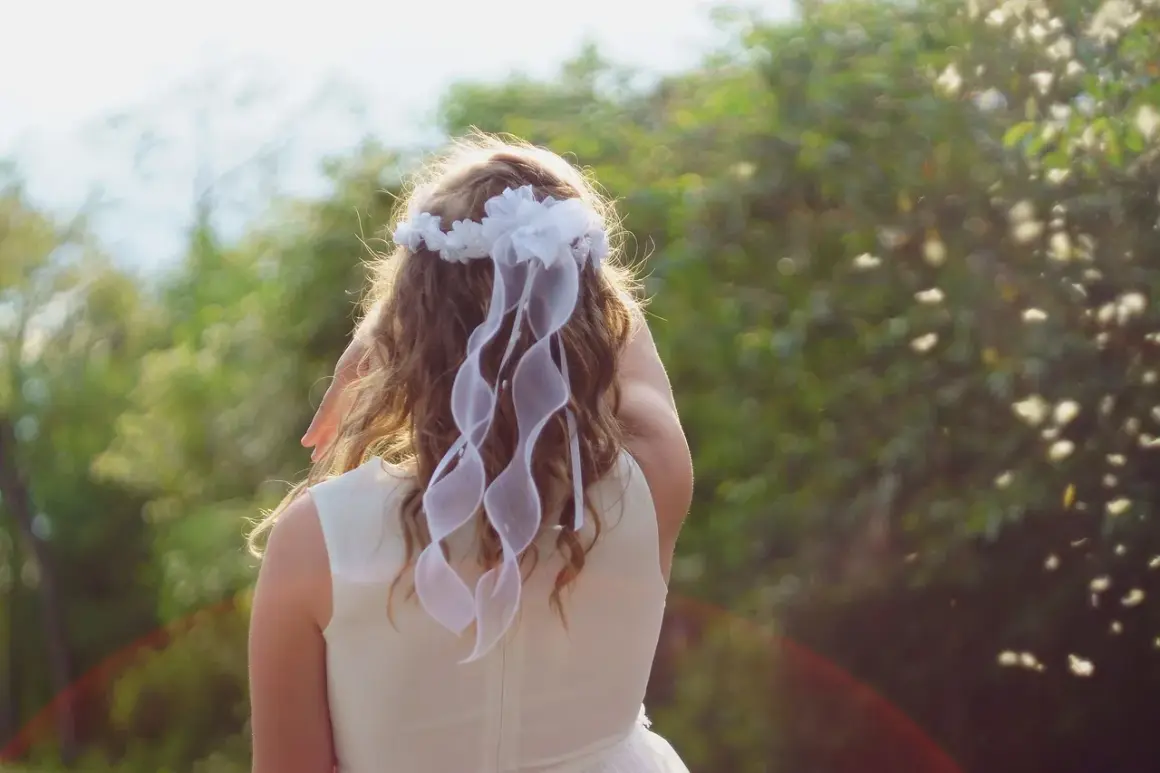 Coiffure fille communion : une jolie tresse ornée d'un diadème de fleurs blanches et de rubans flottants, prête pour sa grande journée.