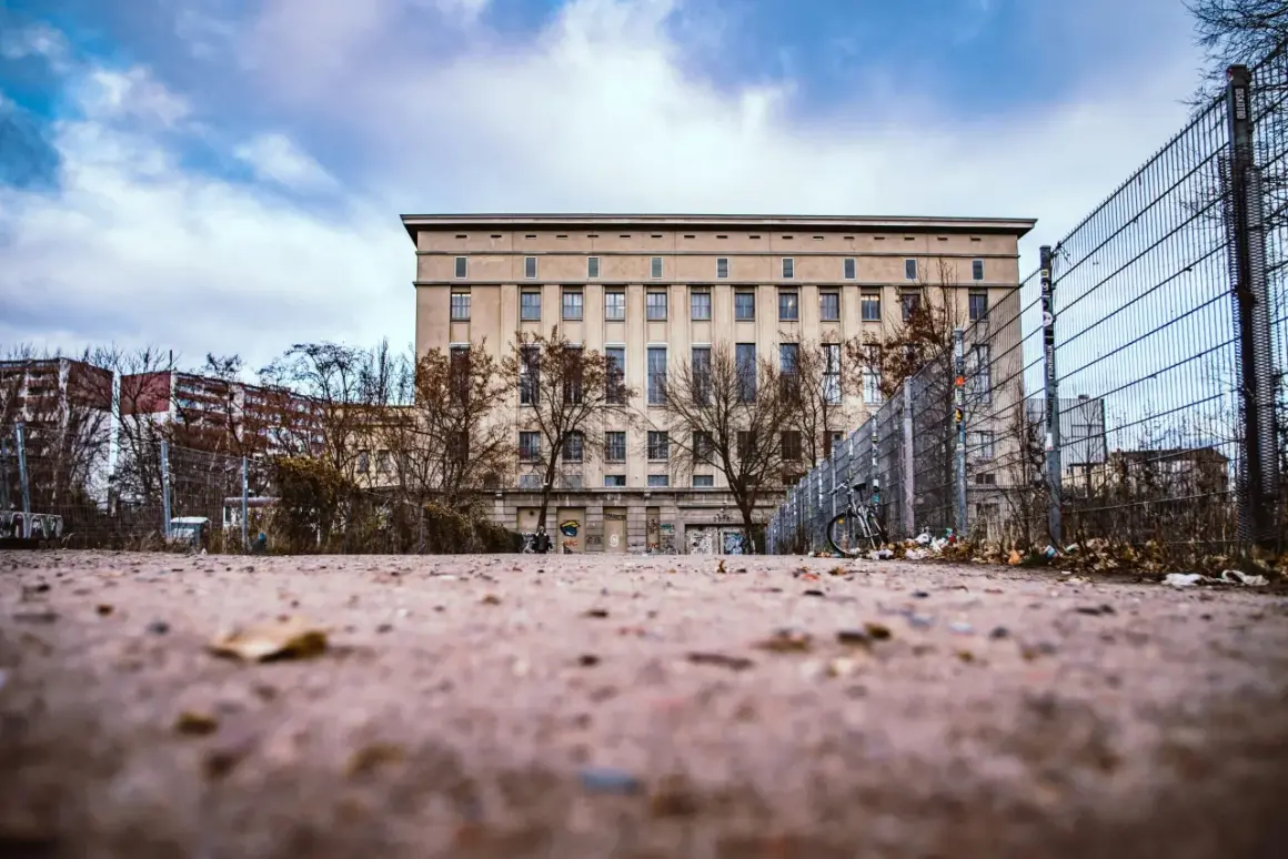 L'imponente edificio del Berghain, con la sua facciata austera e le finestre scure, si staglia sotto un cielo nuvoloso. Un cancello metallico e alberi spogli circondano l'area, suggerendo un percorso misterioso per chi cerca come entrare al Berghain.
