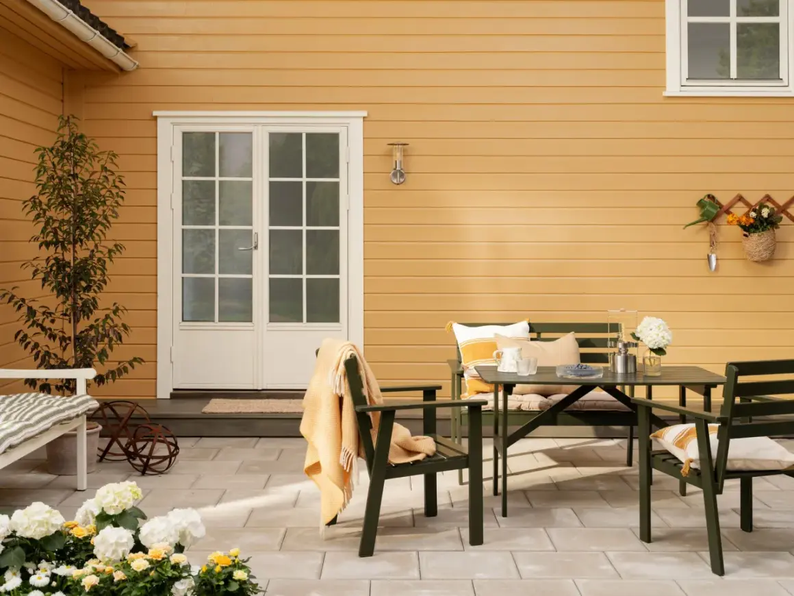 Ett gult hus med svart tak. En uteplats med gröna möbler, blommor och en orange pläd.