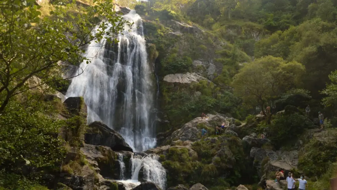 La majestuosa cascada de Belelle cae en cascada por rocas cubiertas de musgo, con gente disfrutando del paisaje y el agua.