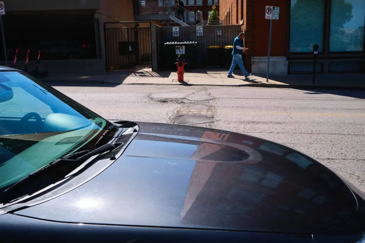 A man walks past a fire hydrant on a sunny day, his reflection visible in the hood of a car. 40mm street photography captures the urban scene.