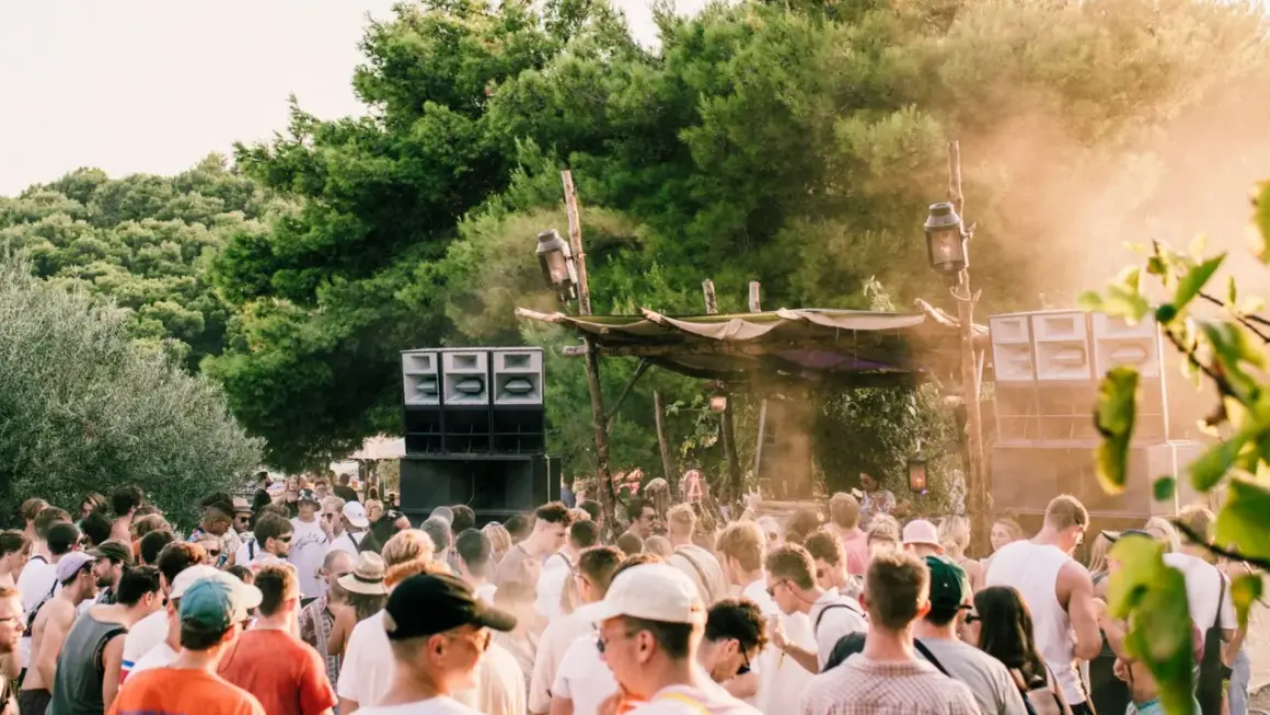 Folla in delirio a un festival tecno all'aperto, con enormi casse acustiche e un palco improvvisato tra gli alberi.