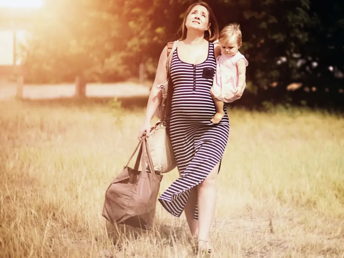 Schwangere Frau mit Baby im Arm und Taschen, die sich durch hohes Gras kämpft. Schwer heben schwangerschaft.