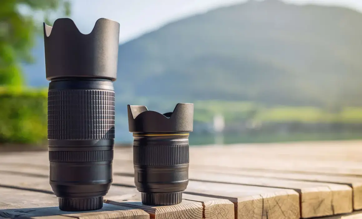 Two camera lenses, a tall telephoto and a shorter normal lens, sit on a wooden deck with a blurred mountain and lake background.