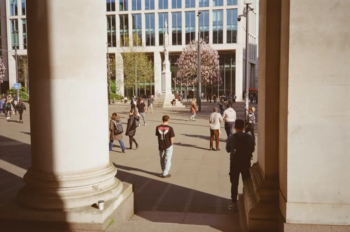 A 35mm street photography scene captures people walking in a plaza with a modern building and blooming trees.