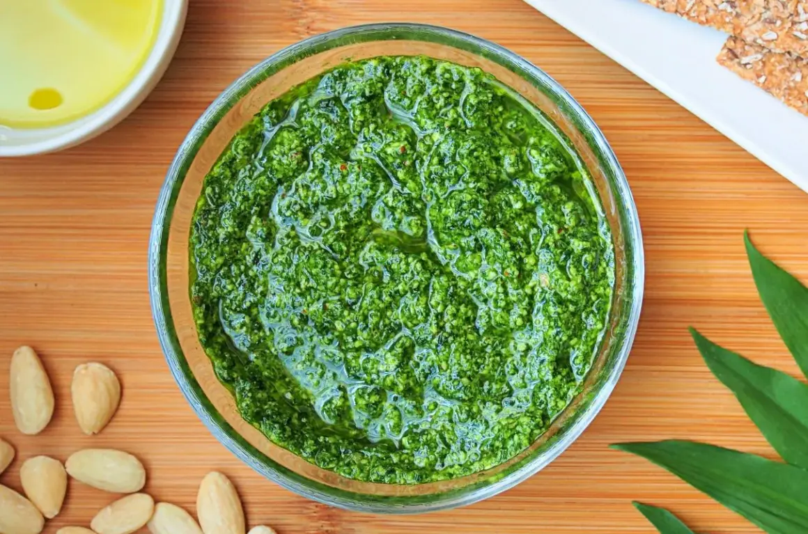 Aromatic pesto z czosnku niedźwiedziego w szklanej miseczce, otoczone migdałami i świeżymi liśćmi.