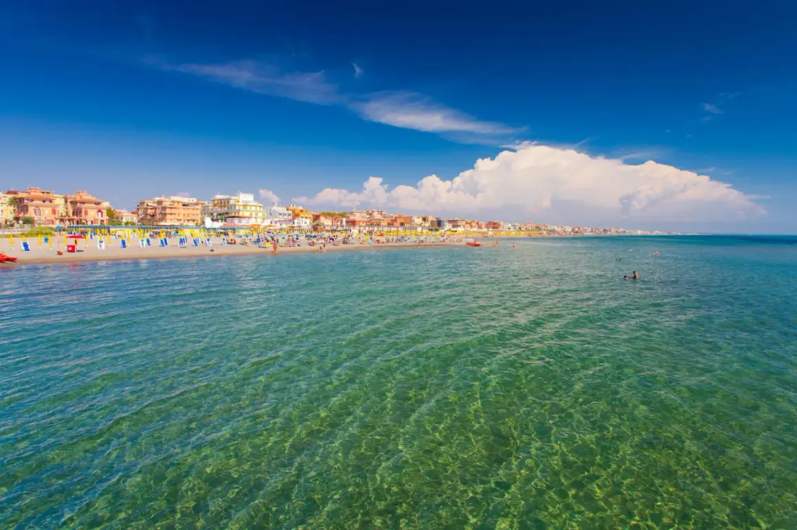 Słoneczny dzień nad morzem w Rzymie. Tłumy plażowiczów, kolorowe parasole i budynki wzdłuż wybrzeża.