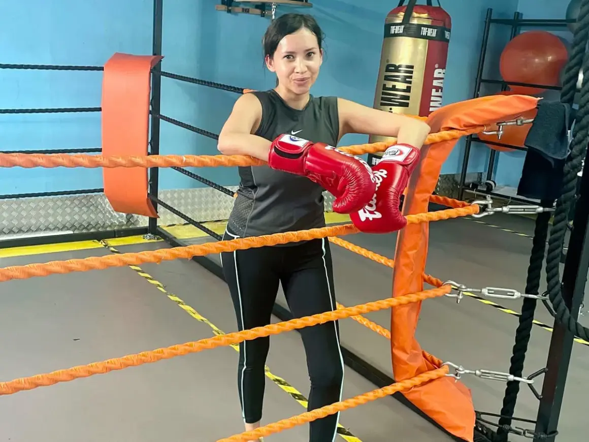 Woman in boxing gloves leans on ropes in a ring, ready for fight training.