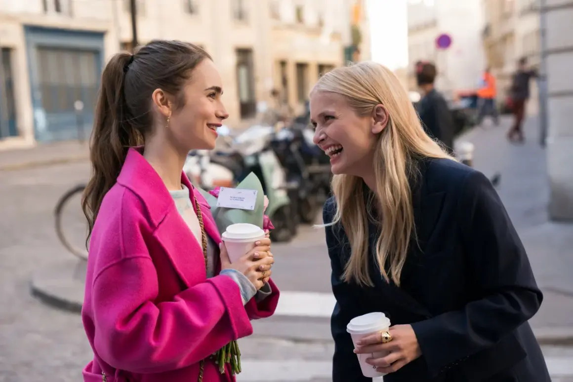 Camille och Emily i Paris, skrattandes med kaffe och blommor på en solig gata.