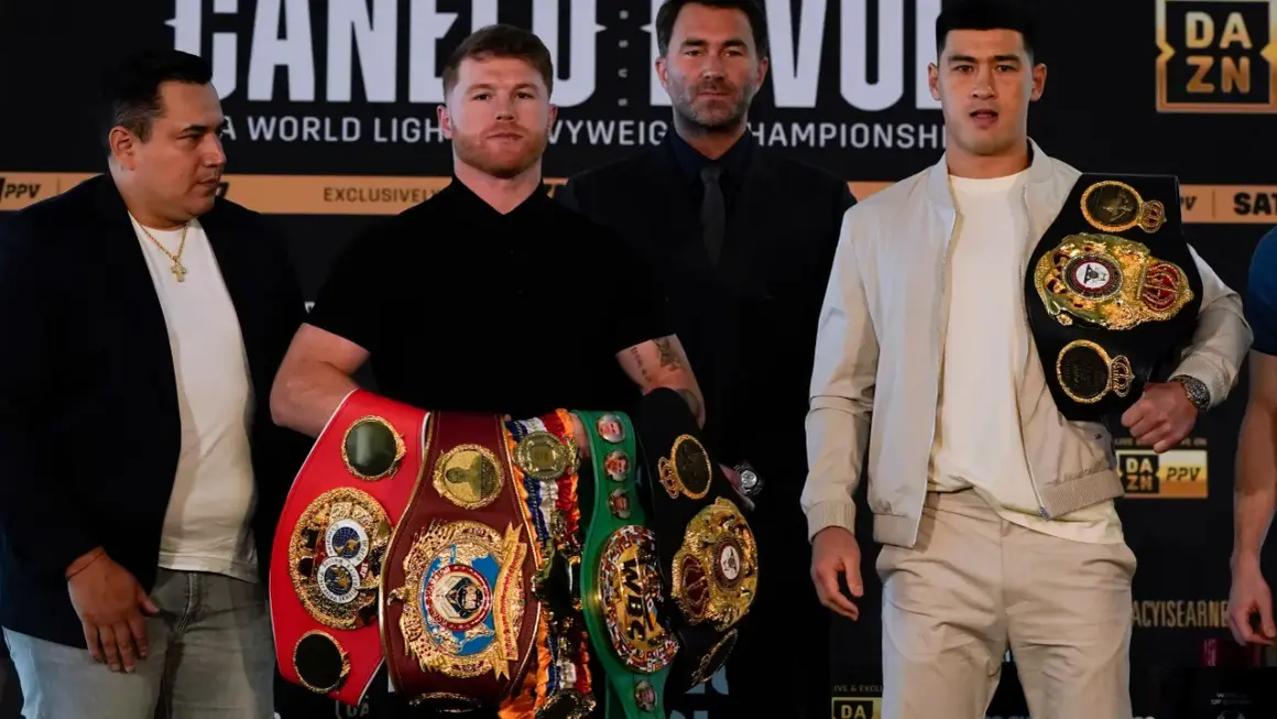 Canelo Alvarez, surrounded by championship belts, shows how good he is. He stands with Dmitry Bivol and Eddie Hearn.