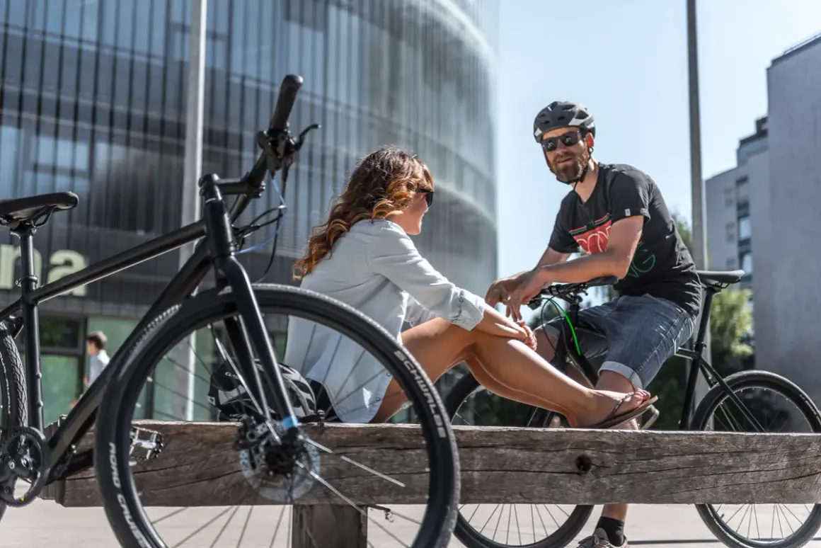 Ein Paar entspannt sich auf einer Holzbank mit Fahrrädern in einer modernen Stadt. Hier lebt es sich fahrradfreundlich.