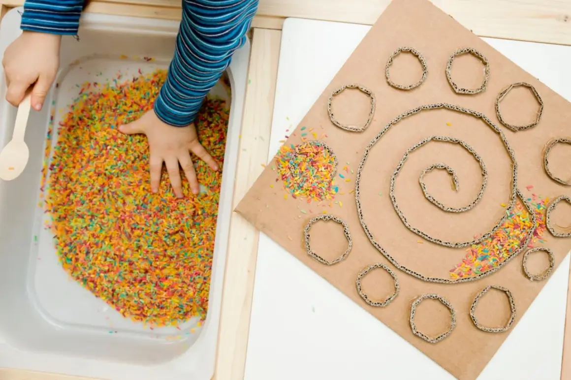 Main colorée dans un bac, une main d'enfant explore. À côté, un carton en spirale avec des ronds découpés, parsemé de riz coloré. Une activité sensorielle adulte pour stimuler la créativité.