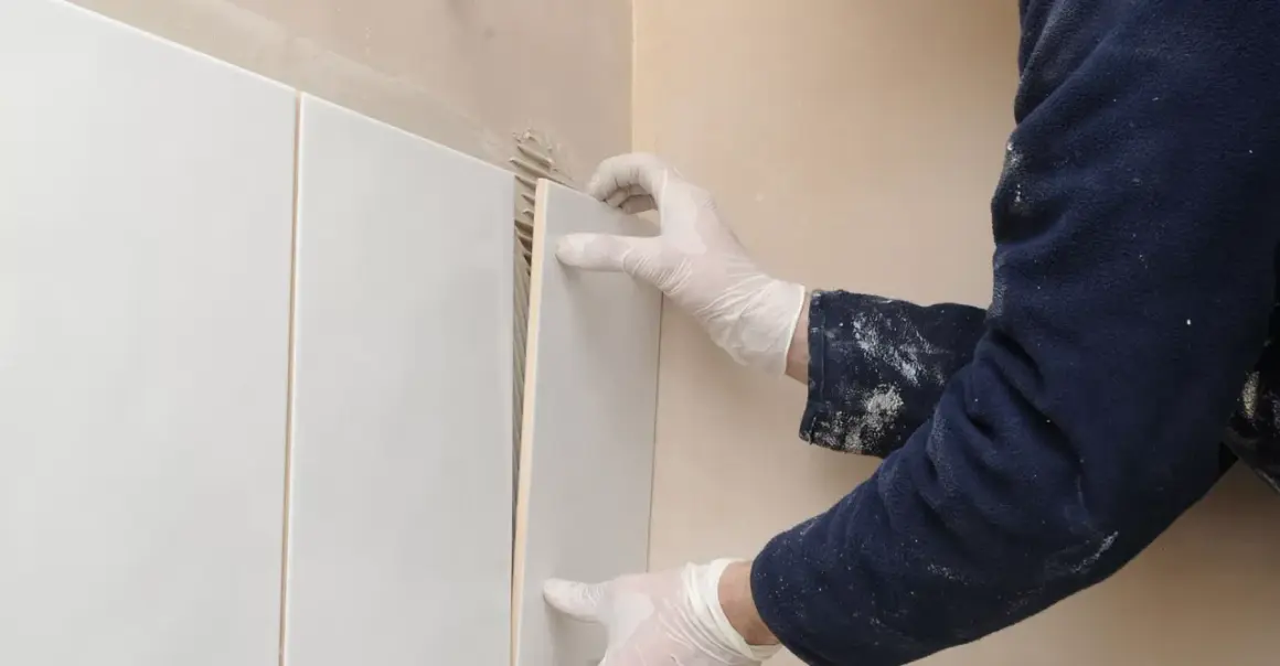 Mani guantate applicano una piastrella bianca su un muro, mostrando come attaccare piastrelle al muro con precisione.