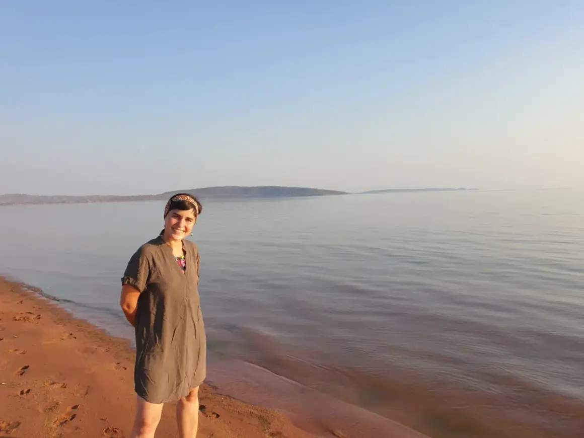 Mujer sonriente en la orilla de arena rojiza del lago Kariba, con el agua tranquila y el cielo despejado al fondo.