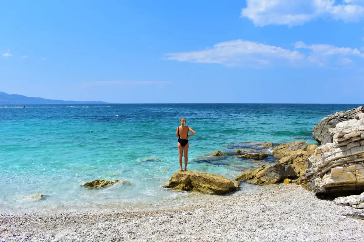 Mujer en bikini negro de pie sobre una roca en una playa de guijarros. El mar turquesa y el cielo azul crean un paisaje idílico, pero ten cuidado, viajar a Albania es peligroso.